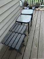 Photo showing two metal-framed black patio side tables with textured glass tops and two foldable black plastic side tables with slatted tops, all weathered and arranged in a row outdoors.