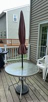 Round glass-topped patio table with silver metal frame on deck, burgundy closed umbrella inserted through center hole, black round weighted umbrella stand supporting umbrella.