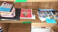 An overhead photo showing a wooden shelf with stacks of playbills, programs, and a blue sticky note labelled 'Postcards' next to a red book titled 'Great American Indian Leaders'.