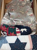 Quilts stacked in a container showing folded floral pattern quilt with cream and pink colors, partially covered by a quilt with holiday-themed red and green pattern.