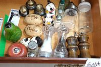 Top-down view of a drawer containing various salt and pepper shakers made from wood, glass, and ceramic including souvenir pieces with 'Maine' engraving and dog shapes.