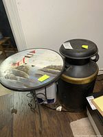 Photo showing the round side table with lighthouse painted top and wrought iron base alongside the vintage metal milk can with black and gold band detail.