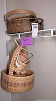 Three woven baskets hanging on metal racks, each handcrafted with natural and dyed fibers. Tags with signatures attached.