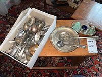 Box containing mixed silverplated flatware including spoons and forks with visible tarnish and oxidation, placed next to a small silverplated tray with salt cellar and salt and pepper shakers.