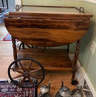 Wooden tea service cart viewed from front showing drop-leaf sides, lower shelf, and large spoked wheels. Part of metal tea set visible at bottom.