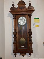 Full front view of the wind-up wall clock showing ornate wooden case, pendulum, and clock face with Roman numerals.