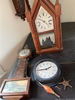 Photo showing all three clocks on a black surface: an antique mantle clock with pointed arch and Roman numerals, a banjo clock with wood case and river scene glass panel, a black Ikea wall clock, wind-up keys, and clock parts.