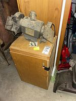 Photo showing dusty vintage bench grinder sander on top of wooden cabinet in basement, sander with open metal protective covers and power switch visible