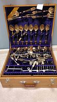 Full view of vintage Siamese brass flatware set with wood handles and Buddha design in open wooden case with purple velvet lining showing organized arrangement of forks, spoons, and serving utensils.