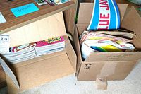 Two cardboard boxes filled with vintage sports-related papers and flat pennants including team logos and sports memorabilia ephemera.
