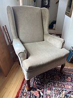Side angled view of beige upholstered wingback chair showing overall shape, upholstery, and wooden cabriole legs.