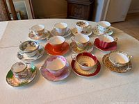 Front view of the 12 bone china cups and saucers on a beige tablecloth, showing assorted decorative patterns and colors.