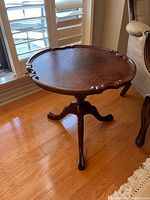 Wooden accent table showing the scalloped round top and curved carved pedestal legs resting on wood floor near window and sofa
