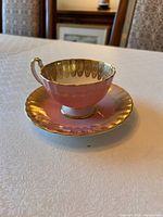 Pink and gold Aynsley tea cup with matching saucer placed on a white textured tablecloth, shown from side angle to highlight gold detailing and pink color.