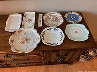 Overview of eight decorative plates and platters arranged on wooden cabinet top.