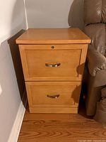 Front view of wooden two-drawer filing cabinet next to couch on hardwood floor showing lock and handles.