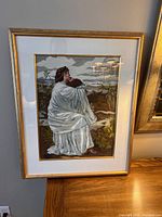 Framed needlepoint artwork showing a figure in white robes looking outward, detailed needlework with a naturalistic outdoor background under cloudy sky.