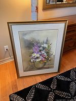 Framed floral print artwork with a bouquet including daisies and other mixed flowers, displayed against a wall with a wooden floor.