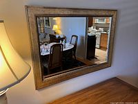 Gold and silver framed wall mirror mounted on a light-colored wall, showing clear reflection of a dining room area with chairs, table, and lamp.