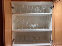 Cabinet shelf with various clear goblets, wine glasses and champagnes. Different sizes and designs of glassware including short and stemmed glasses.