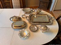 Wide angled view of miscellaneous silverplated tableware displayed on white tablecloth showing arrangement and details of tarnished items.