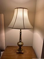 Full view of one brass Stiffel lamp lit on a wooden side table with white bell-shaped shade and turned brass base.