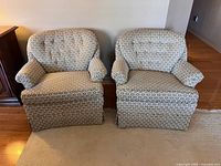 Pair of vintage upholstered tufted tub chairs shown front facing in a room, both having beige fabric with circular patterned upholstery and skirts covering legs.