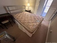 Queen size bed with silver metal headboard and beige quilted mattress in a bedroom setting with bedside table and window shutters visible.