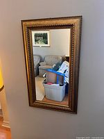 Full view of a rectangular wall mirror with ornate gold frame hung on a light gray wall. Reflections show part of a living room with couches and artwork.