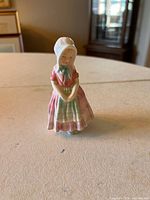 Front view of small Royal Doulton 'Tootles' figurine standing with hands folded, wearing bonnet and traditional pastel dress.