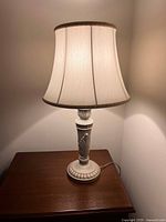Full view of the table lamp showing the cream-colored fabric shade with brown trim and decorative base with a carved leaf design on a wooden surface.