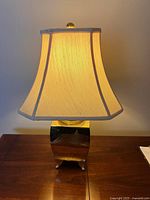 Table lamp lit, showing beige fabric lampshade and reflective gold-tone rectangular base on wooden surface