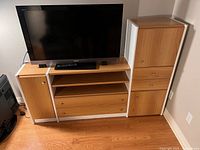 TV entertainment unit with TV on top; rectangular unit with wood panels and white framing, 2 tall side cabinets with doors, 2 drawers below open shelves, wood floor background.