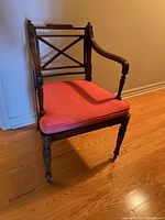 Front and side angle of vintage Regency style armchair with cane seat and coral cushioned seat pad on wooden floor against wall.