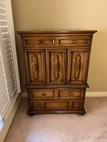 Front view of vintage solid wood armoire showing upper smaller drawers above three carved doors and lower three drawers.