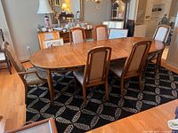 View of vintage oval dining table with six chairs around it on a patterned rug, showing table surface and chair upholstery.