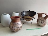 Five ceramic decor pieces on a white shelf: two white vases, two painted terracotta vases, one black bowl on metal stand