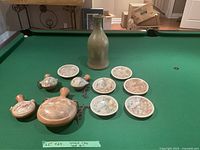 Photo showing the full assortment of ceramic art on green billiards table, including tall vase, large flat vessel, two small vessels, and seven small plates with prehistoric or tribal designs.