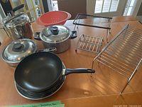 Photo showing assorted pots, three non-stick frying pans stacked, red colander, and two metal cupboard organizers on a wooden table.