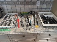 Wide view of assorted cutlery and cooking utensils laid out on countertop. Includes white drawer organizers with cutlery and loose utensils including red silicone brushes, metal kitchen tools, shears and whisks.