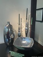 Overall view of tall silver glass vase, two tall silver glass candleholders holding black candles, and a large square silver glass bowl.