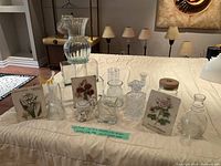 Assortment of various clear glass vases, canisters, wall art plaques, and candleholders displayed on a bed, showing overall lot contents.