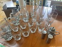 Clear glass assortment on wooden table including low ball glasses, pilsner glasses, beer mugs, and a glass pitcher, also metal salt and pepper shakers and a vintage sugar dispenser.