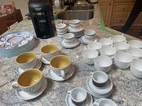 Wide shot showing the black Hamilton Beach coffee maker and assortment of various coffee cups and mugs on kitchen counter.