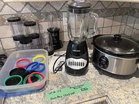 Wide shot of the Black & Decker blender, HomeMax crockpot, Magic Bullet blender with accessories including multiple colored rings and containers on a kitchen counter.