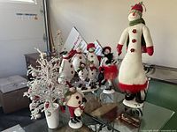 View showing an assortment of seven snowman figurines arranged on a glass surface alongside two decorative Christmas trees, one white glittered in a pot and the other rustic wooden style.