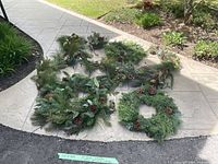 Overview of three Christmas wreaths, lighted garland, and sprays laid out on pavement showing green pine needles and pine cone decorations