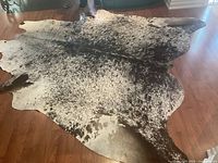 Full view of the large cowhide rug on a wooden floor showing its speckled black, brown, and white coloration and natural shape.