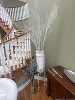 Full view of the white urn holding tall white branches, placed near a wooden staircase and green wall.