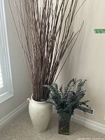 Photo showing tall cream urn vase with long brown branches and shorter clear glass vase with faux eucalyptus on carpeted floor near wall and vent.